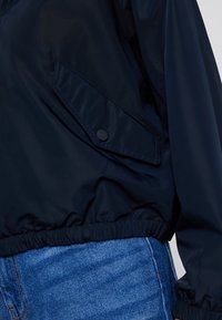 Granatowa bomberka z elastycznym dołem, z boczną kieszenią zapinaną na okrągły zatrzask. Gładki materiał i luźny krój.