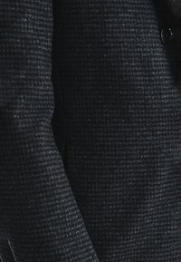 Ciemny, teksturowany płaszcz z subtelnym wzorem w kratkę, z zapięciem na jeden guzik i dopasowanym krojem. Miękka, przypominająca wełnę tkanina.