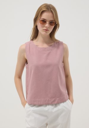 Junge Frau mit Sonnenbrille, ärmellosem staubrosafarbenem Top und weißer Hose, die mit den Händen in den Taschen vor einem einfarbigen Hintergrund steht.
