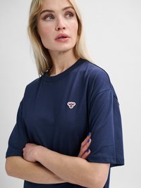 Blonde vrouw met blauwe ogen draagt een marineblauw T-shirt met een klein geborduurd bijenembleem, armen gekruist, kijkt opzij.