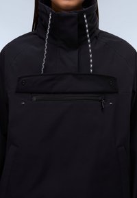 Chaqueta negra de cuello alto con cordones en la capucha y un gran bolsillo en el pecho con cremallera, llevada por una persona de pie.