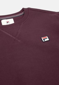 Sudadera burdeos hecha de tejido suave, con cuello redondo acanalado y un pequeño logotipo bordado en la parte superior izquierda.