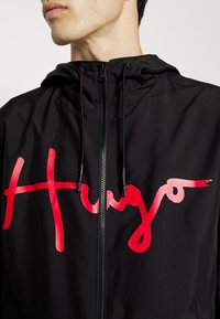 Black hooded jacket featuring a red graphic "Hugo" logo. Smooth, lightweight fabric with a full zipper and adjustable drawstring hood.