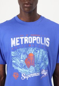 Blaues Baumwoll-T-Shirt mit einem grafischen Design einer Stadtsilhouette und dem Text "METROPOLIS", "DAILY PLANET" und "Superman" in Orange und Weiß.