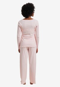 Woman with curly hair wearing light pink long-sleeve top and matching wide-leg pants standing facing away on white background.