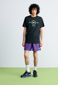 Jovem com cabelo encaracolado, vestindo uma t-shirt preta da Nike, calções roxos da Nike, meias brancas e ténis pretos da Nike, em pé sobre um chão verde.