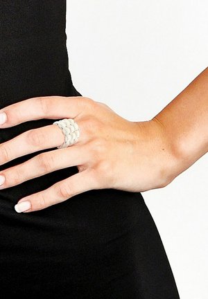 Bague blanche et flexible avec un design en couches présentant des formes arrondies et de petits accents métalliques. Portée sur une main devant un fond noir.