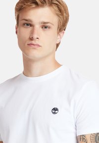 Weißes Baumwoll-T-Shirt mit Rundhalsausschnitt, ausgestattet mit einem kleinen blauen Logo auf der linken Brust. Glatte Textur, Standardpassform.