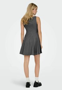 Donna con lunghi capelli biondi, che indossa un vestito grigio scuro senza maniche, calze bianche e mocassini neri spessi, in piedi di spalle.
