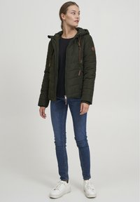 Dark green puffer jacket with a hood, front zip, and drawstrings. Paired with fitted blue jeans and white sneakers. Smooth texture.