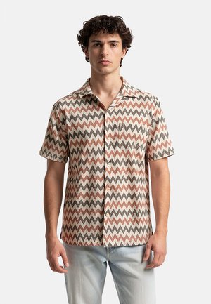 Giovane uomo che indossa una camicia a maniche corte con bottoni, con motivo a zigzag nei colori beige, marrone e ruggine, abbinata a jeans azzurri.