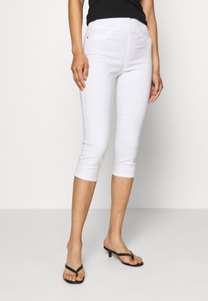 Kobieta ubrana w białe, dopasowane, krótkie spodnie, czarny bezrękawnik i czarne sandały na obcasie na jednolitym tle.