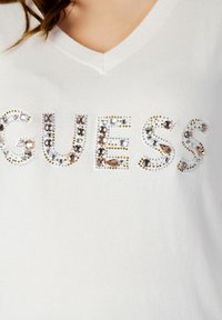 Maglione bianco a V con la parola "GUESS" abbellita da strass colorati e borchie metalliche disposte in un motivo decorativo.