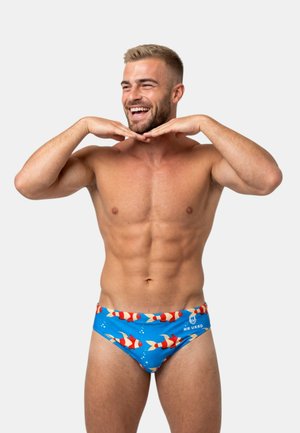 Hymyilevä lihaksikas mies sinisissä uimahousuissa, joissa on punainen kalakuvio, pitää käsiään leuan alla yksiväristä harmaata taustaa vasten.