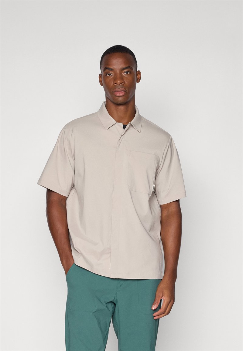 Beige short-sleeved shirt with a collar and a pocket on the left chest. Made of smooth fabric with a relaxed fit. Worn with green pants.
