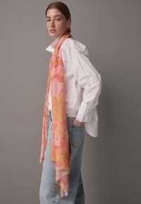 Vaaleanpunainen kukkahuivi, joka on valmistettu kevyestä kankaasta ja jossa on suuret keltaiset ja oranssit kukkakuviot, laskeutuu tyylikkäästi kaulan ympärille.