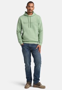 Felpa verde menta con tasca a marsupio e logo tono su tono, abbinata a jeans di denim blu e scarpe marroni, in piedi su uno sfondo bianco.