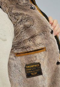 Faux fur jasje in lichtbruin met een gladde textuur, met een binnenzak omlijnd in oranje en een zwart label met "SNOWPACK."