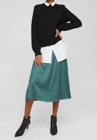Persoon in een zwart sweatshirt over een wit shirt, met een teal midi-rok en zwarte leren enkellaarsjes, staand met één hand op de heup.
