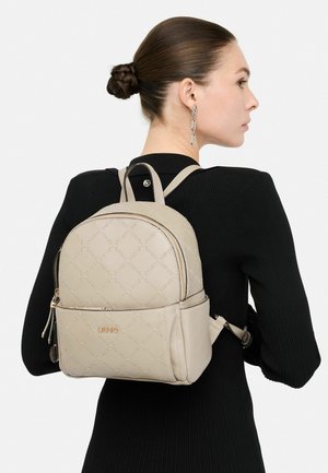 Zaino in finta pelle beige con design trapuntato, dettagli in metallo dorato, manico superiore e spallacci regolabili, indossato sulla schiena.