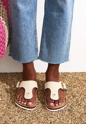 Sandales beiges avec une large lanière et boucle, associées à un jean en denim bleu cropped. Les sandales présentent une semelle plate et une texture en cuir.