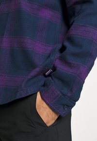 Laivastonsininen ja purppuranvärinen ruudullinen flanellipaita, jossa on napilliset hihansuut ja pehmeä materiaali. Helman lähellä näkyy etiketti.