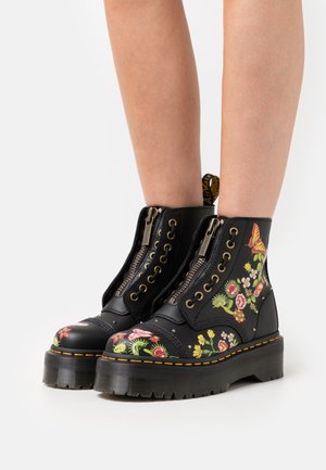Bottines en cuir noir ornées d'une broderie florale colorée, de ferrures dorées, de fermetures éclair et d'une semelle épaisse texturée avec des surpiqûres jaunes.