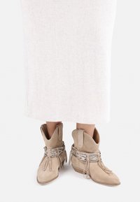 Beige suede ankle boots with pointed toes and tall tops, featuring fringe details and silver hardware accents on the side.
