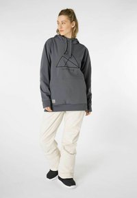 Grauer Kapuzenpullover mit schwarzer Berggrafik und Logo, kombiniert mit cremefarbener Hose. Die Schuhe haben ein schwarzes Obermaterial und eine weiße Sohle.