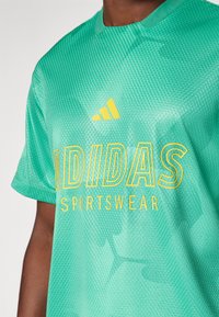 Camisa desportiva verde com acabamento texturizado, com o logo amarelo "ADIDAS" e o texto "SPORTSWEAR" abaixo, e três riscas amarelas no ombro.