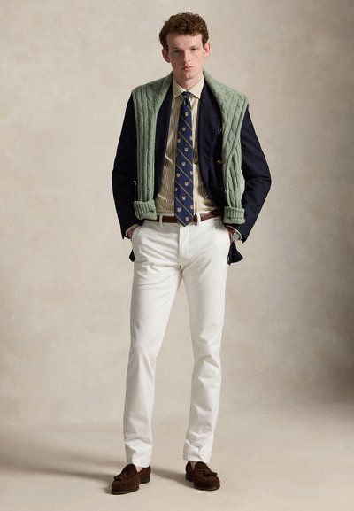 Άνδρας που φοράει μπλε σακάκι, ριγέ πουκάμισο, γραφική γραβάτα, λευκό παντελόνι, καφέ loafers και πράσινο πουλόβερ περασμένο στους ώμους, στέκεται με τα χέρια στις τσέπες.