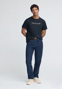 Mörkblå bomullst-shirt med "POLO CLUB"-logotyp, kombinerad med blå denimjeans och ljusbeige sneakers. Outfiten har en avslappnad passform.