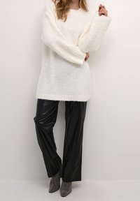 Pull blanc épais à manches larges, associé à un pantalon noir brillant en simili cuir et des bottines métalliques.