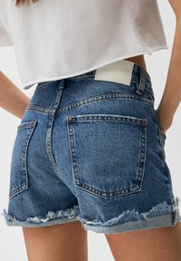 Mujer vistiendo pantalones cortos de mezclilla azul de cintura alta con dobladillos deshilachados y cortos en blanco, con las manos en las caderas.
