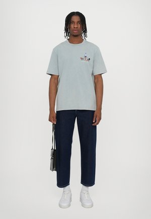 T-shirt en coton bleu clair avec un petit imprimé graphique sur la poitrine, associé à un jean droit bleu foncé et des baskets blanches.