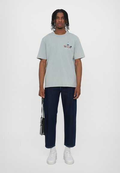 T-shirt en coton bleu clair avec un petit imprimé graphique sur la poitrine, associé à un jean droit bleu foncé et des baskets blanches.
