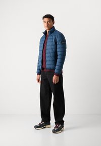 Gevoerd blauwe jas met een hoge kraag, een kastanjebruine gestructureerde trui eronder, zwarte broek en meerkleurige sneakers.