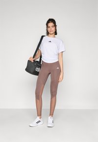 Bijela majica s crnim Adidas logom, smeđe tajice s mrežnim detaljima, crna torba s bijelim logom i bijele atletske tenisice.