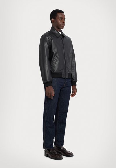 Chaqueta bomber de cuero negro con cuello, cierre frontal y puños de canalé. Llevada con pantalones de mezclilla oscuros y zapatos negros, de pie frente a un fondo blanco.