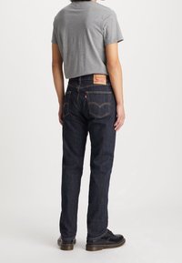 Person som bär mörkblå raka jeans med brun sömnad och svarta skor, står vänd bort mot en enfärgad vit bakgrund.