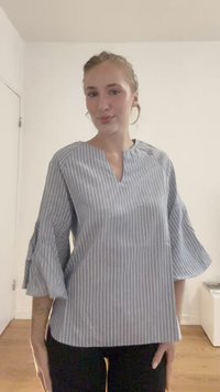 Modra in bela črtasta bluzica z V-izrezom, zvonastimi rokavi in lahkotno teksturirano blago. Nosena z črnimi hlačami.