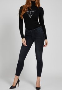 Maglietta nera a maniche lunghe con design del logo, abbinata a jeans skinny in denim scuro. Il modello indossa scarpe con tacco alto nere.