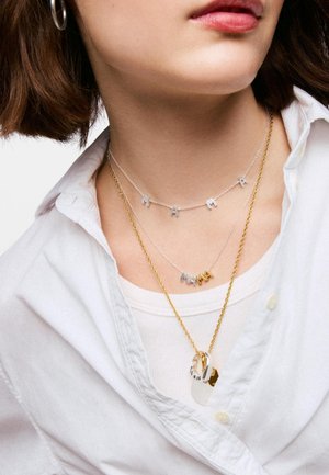 Primer plano de una persona que lleva collares superpuestos, incluyendo cadenas de plata y oro con dijes de letras que forman la palabra "MAMA" y pendientes de aro.