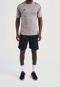 Šedé sportovní tričko s krátkým rukávem a černými akcenty na ramenech, v kombinaci s černými šortkami a černými sportovními botami. Viditelné bílé ponožky.