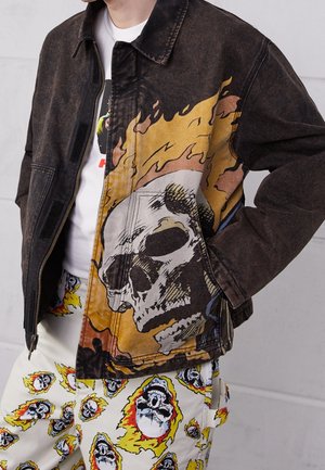 Spijkerjack met een bruinzwarte tint, met een kleurrijk ontwerp van een schedel en vlammen aan één zijde. Gecombineerd met witte broek versierd met schedels en vlammen.