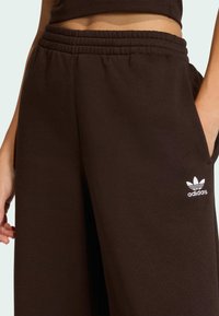 Hnedé športové nohavice z mäkkej látky s elastickým pásom, vybavené bočným vreckom a bielym logom Adidas na spodnej časti nohy.