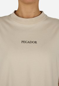 Beigā t-kreklu ar apaļu kakla izgriezumu un īsām piedurknēm, ar tekstu "PEGADOR" melnā krāsā centrā uz krūškurvja. Mīksta tekstūra.