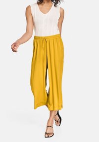 Żółte spodnie o szerokich nogawkach z elastycznym paskiem i bocznymi rozcięciami, zestawione z fakturowanym białym topem bez rękawów oraz czarnymi sandałami na paski.