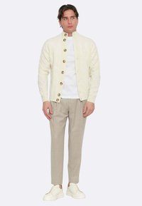 Cardigan bianco lavorato a maglia con motivo a treccia, bottoni marroni, indossato sopra una t-shirt bianca, abbinato a pantaloni grigio chiaro e sneakers bianche.