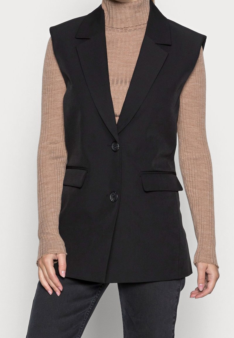Black sleeveless vest with a tailored design, fabric buttons, and two front pockets, worn over a beige ribbed turtleneck.